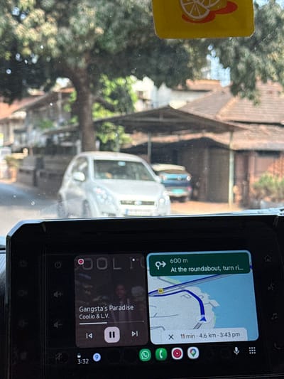 Captured from inside a vehicle, the image presents a street scene in Reis Magos, India. The view is partially obscured by a dirty windshield, featuring a yellow sticker in the upper left. Through the windshield, we see an older home with a tiled roof, along with trees and glimpses of other vehicles on the road. Attached to the dashboard of the vehicle is an infotainment system screen displaying a navigation app indicating that the driver is 600 meters from a roundabout and should turn right. The app estimates the destination is 4.6 kilometers away and will take 11 minutes, arriving at 3:43 pm. The screen also shows the song "Gangsta's Paradise" by Coolio & L.V. is paused and being played at 3:32.