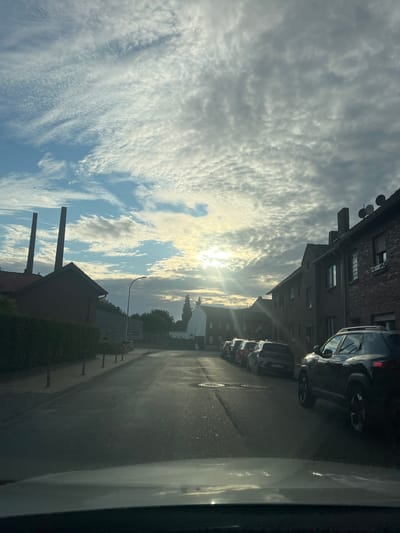 This is a photograph taken from inside a vehicle, likely a car, showing a street scene in Heinsberg, Germany. The image is oriented vertically.

The primary subjects are a row of parked cars and a brick building. The cars are dark in color and appear to be SUVs or similar vehicles, lined up along the asphalt road. The building is made of red brick and has multiple windows. In the background, more buildings and trees are visible. The foreground shows a portion of the interior of the vehicle the photo was taken from, specifically a white surface that reflects the sky and surrounding environment.

The scene depicts a quiet street, possibly in a residential or commercial area. There are no people visible. The activity is static, with cars parked and buildings standing.

Notable details include the time of day, indicated by the bright sunlight streaming through the clouds. The sun is partially obscured by thin, wispy clouds that create a dramatic sky. The clouds are white and grey, suggesting a partly cloudy day. The lighting creates long shadows on the road, further emphasizing the time of day, which appears to be late afternoon or early evening.

There is some text visible on the license plate of one of the cars, but it is illegible. The overall mood of the image is peaceful and somewhat serene, due to the soft lighting and the quiet street.