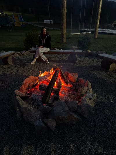 An outdoor night scene depicts a solitary female subject seated on a rustic wooden bench around a bonfire within the Paracin Municipality, Serbia. The bonfire, centrally located in the foreground, is contained by a ring of irregular grey stones and exhibits actively burning wood, embers, and ash, emitting orange and red light. The ground surrounding the fire and benches is gravel.
The female, a young adult, is positioned behind the bonfire, facing forward with a slight turn to her left. She is dressed in a dark jacket, light-colored sweatpants, and sneakers. Her left hand holds what appears to be a mobile phone, while her right hand rests on her lap. A silver-colored can rests on the bench to her right. She exhibits a smiling expression.
Two other rustic wooden benches, similar in construction to the one occupied, are partially visible to the far left and right of the gravel area. In the background, beyond a grassy expanse, tall trees are visible, one featuring a swing set. To the far left, a yellow and blue children's slide structure is present. A white, tent-like structure is discernible in the distant background. The sky is dark, consistent with nighttime conditions. The environment suggests a recreational or glamping site.