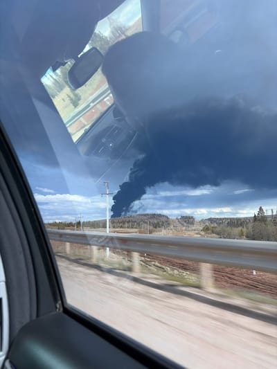 The image captures an outdoor scene from the interior of a moving vehicle, presented in a portrait orientation that has been rotated 90 degrees clockwise. The primary subject is a large, dense plume of black smoke rising dramatically from the ground on what would be the left side of the correctly oriented image, extending upwards into a dark, overcast sky.

In the mid-ground, a highway or road, blurred by motion, runs parallel to a metal guardrail. Beyond the road, a forested landscape or hilly terrain is visible under a sky that transitions from clear blue with scattered white clouds on the right to heavy, dark clouds on the left, coinciding with the smoke plume. Several utility poles stand alongside the road.

The foreground of the image features reflections on the car's windshield or sunroof. These reflections include elements of the vehicle's interior, such as a portion of the dashboard, air vents, and a rearview mirror. Faint reflections of distant buildings or structures can also be seen.

The activity or event taking place is a significant fire or industrial incident, evidenced by the large column of black smoke. The perspective suggests the photographer is observing this event while traveling along a road. The scene is set during daytime, likely late morning or afternoon, given the natural light. The weather is mixed, with bright skies contrasting sharply with the ominous, dark clouds associated with the smoke. The location is Kliuchiki (Kultaevskoe S/P), Russia. No visible text is discernible in the image.