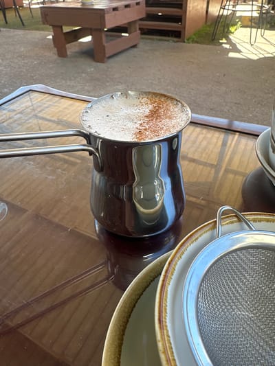 The image shows a close-up of a coffee cezve filled with frothy coffee, dusted with cinnamon, resting on a glossy, dark brown table. The cezve is made of stainless steel with two handles. In the foreground, a stack of plates and a fine-mesh sieve are visible. The background suggests an outdoor setting, possibly a cafe, with a wooden bench and some greenery, hinting at a location in Bellingen, Australia. The lighting indicates it is daytime, with sunlight casting shadows.