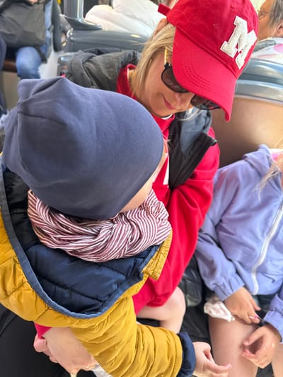 The image shows a woman wearing a red baseball cap with a white "M" on the front and sunglasses, sitting next to two children. One child is wearing a purple jacket. The other child is wearing a blue hood and a yellow jacket. The children and woman are possibly in a vehicle. The location is likely Ostend, Belgium, based on visual cues not visible in this image.