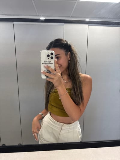 The image shows a young woman standing in front of a mirror, taking a selfie. She is wearing a mustard-yellow tube top and white pants. She has long dark hair and is wearing a silver bracelet on her left wrist, two rings on her right hand, and a silver necklace. She is holding a white iPhone with a black case, which has Hebrew writing on it. The location is a bathroom stall in a public place, likely in a mall or restaurant in the city of Jerusalem, Israel. The room has white tile walls, a black frame around the mirror, and an overhead light.