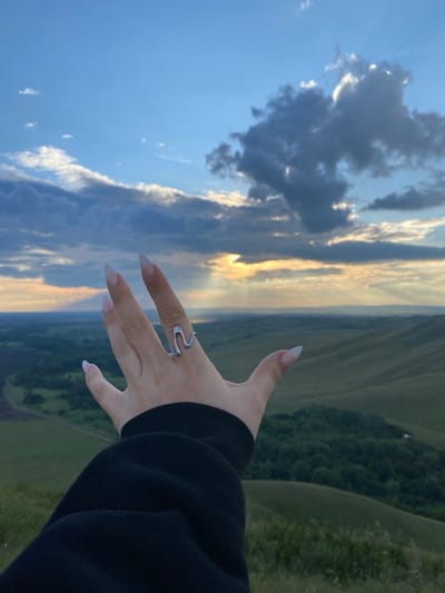 A hand adorned with a silver snake-shaped ring is extended towards a vast, undulating landscape under a dramatic sky. The hand, with long, almond-shaped nails, is positioned in the foreground, partially obscuring the view of the distant terrain. The landscape is characterized by rolling green hills, some sparsely covered with trees and others appearing as open fields. A winding road is faintly visible in the mid-ground. The sky is a blend of deep blue and scattered clouds, with the sun breaking through in a powerful display of crepuscular rays, illuminating the horizon. The overall impression is one of serene natural beauty, with a touch of artistic flair from the ring and the hand's pose. The location is identified as Абзаново, Russia.
