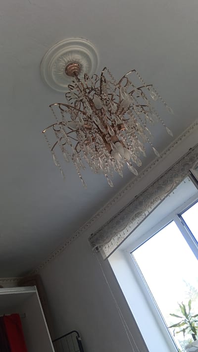 A crystal chandelier with a copper-colored frame hangs from a white ceiling adorned with decorative molding. The chandelier is multi-tiered and features numerous glass crystals that catch the light. Below the chandelier, a portion of a window is visible, with a white frame and sheer lace curtains. The window offers a view of greenery outside, suggesting an outdoor environment. In the lower left corner of the image, a white shelving unit with some red and brown fabric items is partially visible. A metal rack is also present on the floor. The overall scene depicts an indoor residential setting. There are no people visible in the image. The location of Maqat, Kazakhstan is not discernible from the visual information in the image.