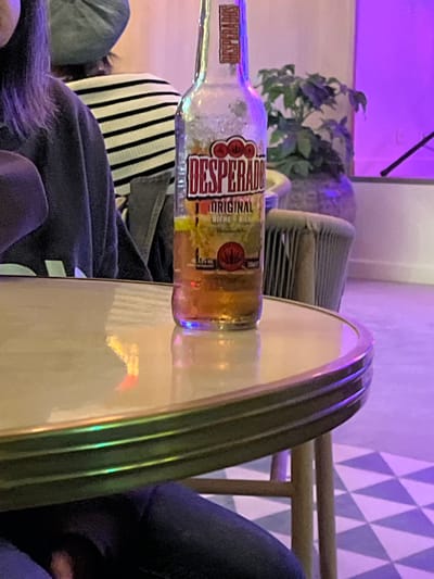 The image shows a Desperados Original beer bottle resting on a light-colored, round surface, possibly a table. A person wearing a dark-colored, possibly navy, sweater is partially visible in the foreground. A black-and-white striped object, potentially a cushion or part of clothing, is located beneath the bottle. In the background, there is a potted plant, a blurred gray object (possibly another piece of furniture), and a wall with a geometric triangle pattern. A faint purple light washes over the background elements. There is no information suggesting the location of Grimaud, France in the image.