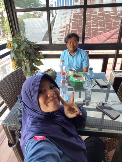 Two individuals, a woman in a purple hijab and a man in a blue polo shirt, are seated at a glass-topped table in an outdoor seating area. The woman is in the foreground, taking a selfie with her right hand raised in a peace sign. She is wearing a blue shirt with a red and white logo patch on the left sleeve. The man is seated behind her, looking towards the camera with a neutral expression. On the table are two bottles of water, a cup on a saucer, a small cardboard container, and two smartphones. The background shows a cityscape with buildings and trees, suggesting an urban or suburban location. The overall scene appears to be a casual meeting or meal at a cafe or restaurant in the city of Keraton, Indonesia.
