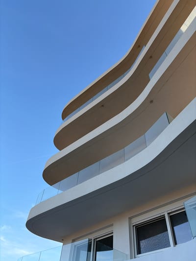 The image shows a low-angle view of a modern building featuring curved architectural elements. The building is characterized by a series of horizontal, undulating shapes that create a sense of depth and movement against a clear blue sky. On the right side of the frame, there is a section of the building with windows and glass balconies, suggesting a residential or commercial structure. The overall style is contemporary, with clean lines and a minimalist aesthetic. The location is likely in the Municipal Unit of Elliniko, Greece, based on the architectural style and known developments in the area.