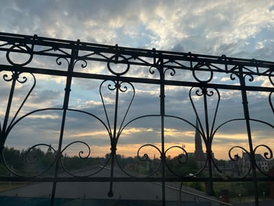 The image shows a view through a decorative metal fence of a cityscape at sunset. The fence is black and ornate, with circular and scrolling designs. In the distance, a tall, red-brick church spire is visible, along with other buildings and trees. The sky is filled with dramatic clouds, illuminated by the warm glow of the setting sun, casting a golden light across the scene. The image appears to be taken from a balcony or window, looking out over the rooftops of Mysłowice, Poland.