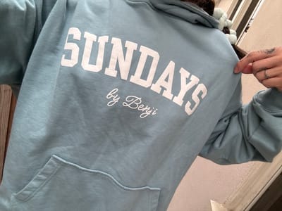 A close-up, slightly angled shot focuses on a light blue sweatshirt. The word "SUNDAYS" is prominently printed in white, block letters on the garment. Above "SUNDAYS," the text "by Berri" is written in a cursive font, also in white. The sweatshirt appears to have a pocket, visible at the top of the image. A hand with a silver ring and some tattoo ink on the wrist is visible on the left side of the image, holding onto the sweatshirt. The background is out of focus, but shows a white wall and what seems to be part of a wooden structure. It is not possible to determine the location in Seal Beach, United States of America from this image.