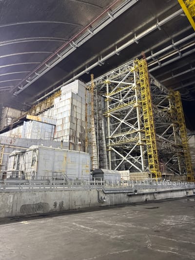 The image depicts a large, industrial structure with a complex metal framework and weathered, paneled buildings. The scene appears to be indoors, under a large, arched ceiling with a dark, textured surface. A prominent yellow metal lattice structure rises on the right side of the frame, interwoven with white beams and platforms. To the left, a series of gray, box-like structures with visible seams and rust are stacked and connected. Metal walkways, railings, and pipes are visible throughout the scene, suggesting a functional, possibly operational, industrial environment. The ground in the foreground is a dark, textured concrete surface, with some scattered lighter patches and what appear to be puddles or damp areas. The lighting is dim and appears to be artificial.

This image was taken at the Chernobyl Nuclear Power Plant in Kyiv Oblast, Ukraine. Specifically, it shows part of the New Safe Confinement structure, an arch-shaped shelter built over the destroyed fourth reactor unit of the Chernobyl power plant.