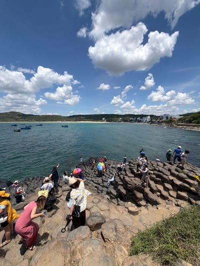 The image shows a coastal scene in Phú Lương, Vietnam, featuring the unique basalt columns of Gành Đá Dĩa, a geological formation. The foreground is dominated by these hexagonal rock formations, with several people navigating the uneven terrain. Some individuals are dressed in casual summer clothing, while others wear conical hats, suggesting a tourist excursion. A few people are holding umbrellas and bags, and some are taking photographs.
In the middle ground, the turquoise ocean stretches towards a sandy beach lined with lush green hills and scattered buildings. Several small blue boats are anchored in the water closer to the shore. The sky above is a vibrant blue, dotted with large, puffy white clouds. The overall impression is a sunny, lively day at a popular natural attraction.