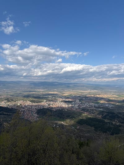 The image presents a high-angle, aerial view of a landscape featuring a town nestled in a valley. On the left side of the frame, a blue sky is visible with scattered cumulus clouds. To the right, the town's buildings are tightly packed, showing a mix of white and red roofs. The valley floor contains agricultural fields of varying colors, indicating different crops or stages of growth. The topography is hilly, with forested areas and rocky terrains visible. In the foreground, foliage from trees or bushes is seen, partially obscuring the view. The town is likely Delchevo, Bulgaria.