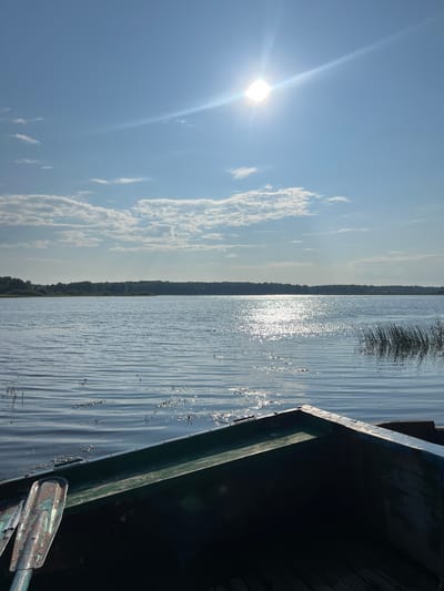 The image depicts a serene lake scene under a bright, sunny sky with scattered clouds. The sun is positioned high in the sky, creating a brilliant reflection on the water's surface. In the foreground, the interior of a dark-colored rowboat is visible, with an oar lying inside. The lake stretches out to a tree-lined horizon, suggesting a natural, possibly rural setting. There are no people visible in the image. The environment is calm and tranquil. Based on the provided information, this scene is located in the city of Kondas, Russia.