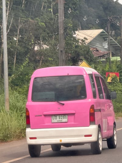 A bright pink van with a white rear bumper and roof is driving on a paved road. The van has a license plate that reads "บจ 9288" and has Thai characters below it. The van is seen from the rear, with the left side visible. The environment appears to be a rural or semi-rural area with lush green vegetation, trees, and utility poles on the side of the road. In the background, there are buildings with sloped roofs and a distant hillside covered in trees. A yellow triangular sign is visible to the left of the van. There are no people or animals visible in the image. The lighting suggests it is daytime. The location is identified as Ban Thung, Thailand.