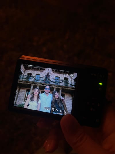 A digital camera shows a photo on its screen. The photo depicts three people in front of a building adorned with Christmas decorations. A man in blue sunglasses stands between two women. The date "2026 12-24 15:53" is displayed on the screen. The camera is being held in someone's hand. A green light is visible on the camera's top surface. The background is dark and blurry.