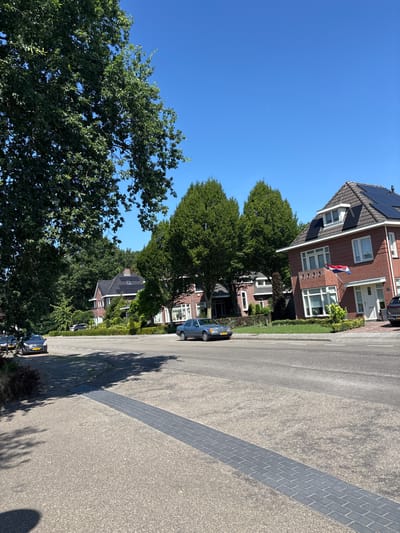 The image depicts a street scene in Asten, Netherlands, on a clear, sunny day. In the foreground, a paved road runs horizontally across the frame, with a patterned pedestrian crossing made of dark gray bricks intersecting it diagonally. The road is lined with parked cars on the left side. Beyond the road, there is a residential area featuring houses with red brick exteriors, dark tiled roofs, and solar panels. Lush green trees and manicured gardens surround the properties. One house displays a Dutch flag. The sky is a bright, cloudless blue, with a faint contrail visible in the distance. The overall impression is one of a peaceful suburban environment.