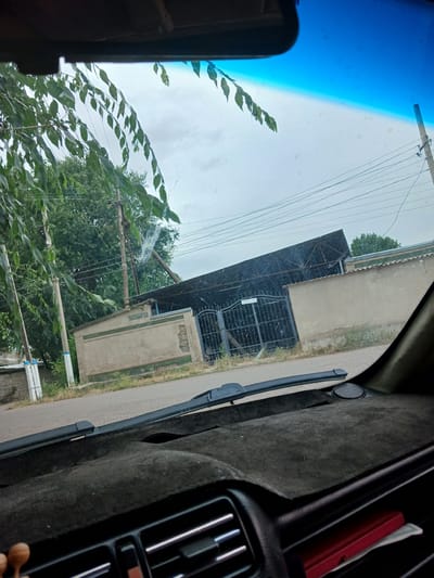 The image captures a view from inside a vehicle, looking out through the windshield. In the foreground, the dark, textured dashboard and a windshield wiper are visible. Beyond the windshield, a street is visible, bordered by vegetation. On the right side of the street, a light-colored, rectangular building with a dark, corrugated metal roof structure attached to its front is prominent. This building has a gate. Further back, trees and utility poles with wires are seen against an overcast sky. The location is identified as Oytal Rural District, Kazakhstan.
