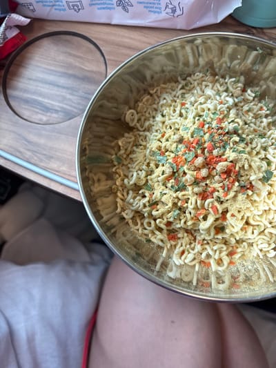 The image depicts an overhead view of a silver metallic bowl filled with uncooked instant noodles and seasoning, resting on a wooden-patterned fold-down tray table. The scene is located within a passenger train, likely in or passing through Tatarsk, Russia, as indicated by Cyrillic text visible on a white package or leaflet in the background.
The instant noodles are pale yellow and have a characteristic wavy, dehydrated form. They are generously covered with a dry seasoning mix comprising yellow powder, small green flakes (likely dried herbs), and orange-red flakes (possibly dried carrots or bell peppers), along with some light beige granular pieces.
The tray table features a prominent circular indentation, typically for holding a cup or bottle. A thin, grey metallic bar is visible running horizontally along the left edge of the tray. In the upper background, a white rectangular object displays various icons and Cyrillic text, including phrases such as "уход за руками" (hand care) and "содержит витамины" (contains vitamins), suggesting a personal hygiene product or informational material.
The lower portion of the image reveals the upper thighs of a person, indicating a first-person perspective. One leg is partially covered by light-colored fabric, while the other is bare, showing skin. A small strip of red fabric is visible between the legs, suggesting part of the person's clothing. No direct actions or interactions are depicted, only the static arrangement of objects. The environment is consistent with a train car interior during a journey.