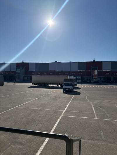 In Novogorelovo, Russia, a large industrial building with numbered loading docks (F-2 through F-8 and G-1 through G-6) is visible under a bright, sunny sky. The building features sections of grey, red, and white panels. A prominent "2F" sign is affixed to a central vertical structure. In the foreground, an asphalt lot is marked with white lines, indicating parking spaces or traffic lanes. Several vehicles are present, including a large white semi-trailer truck with a silver trailer, and a smaller white delivery van. A lone figure, appearing to be a person walking, is observed near the building. A metal railing is partially visible in the lower foreground. The sun is high in the sky, casting long shadows and creating lens flare in the image.