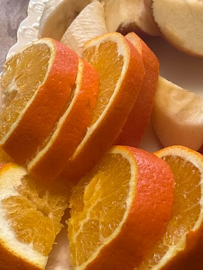 The image shows a close-up of sliced oranges and what appear to be slices of apple or pear on a white plate. The fruits are arranged in a visually appealing manner. The oranges are cut into thin, round slices with visible pulp and rind. The apples or pears are pale yellow and appear to be cut into wedges. The background is blurred, but suggests a textured surface, possibly a tablecloth. There are no people or animals visible in the image. The lighting is bright, highlighting the vibrant colors of the fruit. The image does not provide any information about the location in the city of Getk, Armenia.