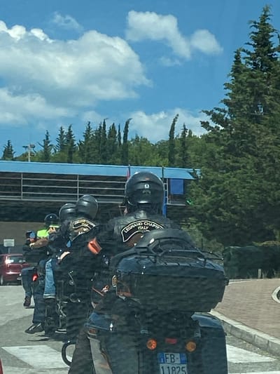 A group of motorcyclists, some wearing black jackets with patches reading "LEGNANO CHAPTER ITALY", are stopped or moving slowly. The motorcyclists are on a paved road, with a red car visible in the background on the left. Trees and a blue structure are visible behind them. The sky is blue with some white clouds. The specific location within Palje Brdo, Croatia is not identifiable from the image.