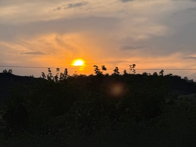 The image depicts a sunset over a rural landscape, likely in Seleznivka, Ukraine. The sun is low on the horizon, casting a warm orange glow across the sky, which is filled with a mix of light and dark clouds. Silhouetted trees and a hill form the foreground and middle ground, with a power line running horizontally across the image. The overall impression is one of natural beauty and tranquility, though the dark foreground suggests the late hour.