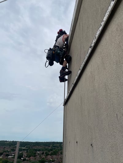 A worker is suspended by ropes along the underside of a concrete overpass. They wear safety gear, including a harness and helmet, and are positioned to work on a metal strip attached to the concrete. Below the overpass, residential housing is visible. The photograph was taken in the Hertsmere region of the United Kingdom under a cloudy sky.