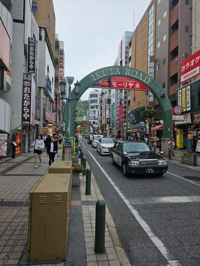 The image is a street scene taken in Kobe, Japan, featuring the “Ikuta Road” archway. The street is lined with shops and restaurants, as evidenced by numerous signs written in Japanese.
A black taxi is driving down the street. Several other vehicles are visible further down the road.
On the sidewalk, two individuals are present. One person is wearing a light-colored jacket and black shorts and appears to be walking. The other person is wearing a black and white outfit.
Other details include: various signs indicating shops and restaurants; green bollards lining the street; buildings with light-colored facades; the “Ikuta Road” arch with signage; utility boxes on the sidewalk.