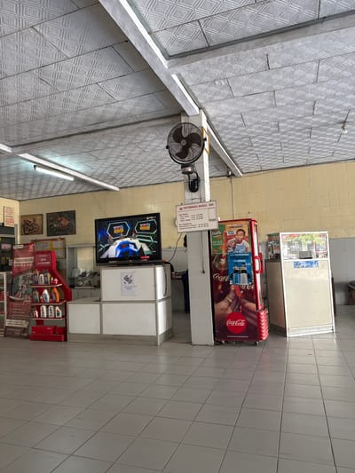 The image depicts the interior of a Honda AHASS motorcycle repair shop in Kebumen, Indonesia. The scene is brightly lit with fluorescent lights and features a tiled floor and a textured ceiling. In the foreground, a red display rack filled with various motor oil bottles is visible. To the right of the display rack is a white counter with a flat-screen television mounted on top, showing a sports broadcast. Next to the counter is a red Coca-Cola branded refrigerator. Further to the right, a cream-colored cabinet with a glass display case stands. The walls are painted in a light yellow hue, and two framed motorcycle posters hang on the left wall. A ceiling fan is mounted above a sign that provides shop information. No people are clearly visible in the frame.