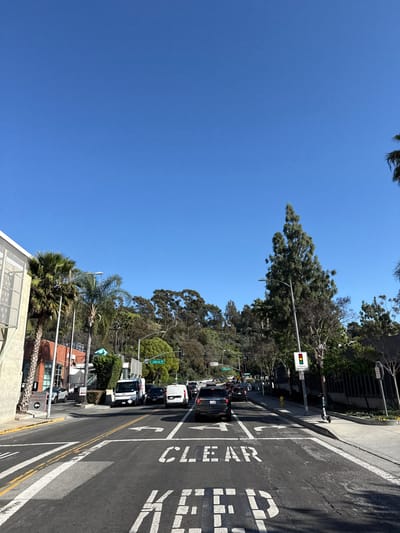 The image depicts a daytime scene at an urban intersection in Culver City, United States of America, specifically at the intersection of Jefferson Boulevard and La Cienega Boulevard. The sky is clear and blue, indicating sunny weather.
In the immediate foreground, an asphalt roadway dominates the lower half of the frame. White road markings include a "KEEP CLEAR" box, with "CLEAR" fully visible and "KEEP" partially visible. Lane division lines and arrows indicate a left-turn lane and a combined straight-ahead/right-turn lane.
Traffic occupies multiple lanes, appearing to be stopped or moving slowly. Vehicles include a dark-colored SUV in the foreground, a white delivery van, and a white United States Postal Service (USPS) truck on the left side, along with numerous other cars extending into the midground. A green traffic light is visible on the left side of the intersection, while a red traffic light is visible on the right side.
To the left of the roadway, a multi-story building with a light-colored, possibly stucco or concrete exterior featuring a grid pattern is visible. Signs near this building indicate "EMERGENCY ENTRANCE" and "PATIENT ENTRANCE" with directional arrows. Several tall palm trees are present on the left, alongside other assorted green foliage.
To the right of the roadway, a paved sidewalk runs parallel to the street. A street lamp post is prominent, attached to which is a traffic signal displaying a red light and a sign for a "PEDESTRIAN ENTRANCE." An electric scooter is parked upright on the sidewalk near the curb. Various trees, including a tall evergreen conifer and several palm trees, line the right side of the street.
The background features a prominent, densely vegetated hill, part of the Baldwin Hills, covered with numerous trees and shrubs under the bright blue sky. No individual people are distinctly visible, but vehicles imply occupants.
