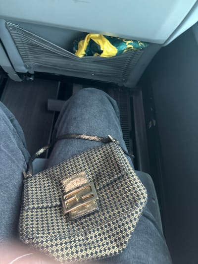 The image shows a person's legs and a purse in a car. The person is wearing dark blue jeans and a patterned purse sits on their lap. The purse is black with gold-colored details, including a large gold-colored buckle with a letter "F" embossed in it. The purse is resting on the person's right thigh. To the left of the image, there is a metal ventilation grate with a green and yellow bag stuffed into it. The image is likely taken inside a car in Otero de Herreros, Spain.  The location was determined using the provided latitude and longitude coordinates.