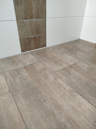The image displays a close-up view of a tiled floor and wall junction. The floor is covered in large, rectangular tiles with a wood-grain pattern in various shades of light brown and gray. The tiles are laid in a staggered pattern. The wall is tiled with large, rectangular white tiles and some tiles with a wood-grain pattern similar to the floor. The wall tiles are arranged in horizontal rows, with some angled to create a decorative effect. A thin white caulk line runs along the base of the wall where it meets the floor. The lighting is neutral, and there are no visible people or other objects in the scene. The image appears to have been taken indoors, likely in a residential or commercial building. The specific location in Kitzingen, Germany, cannot be determined from the visual information provided.