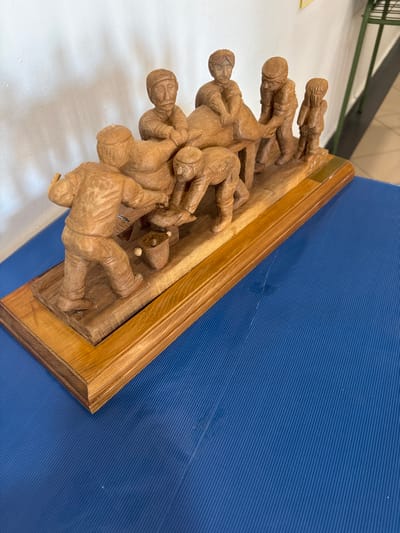 A carved wooden sculpture depicting a group of seven human figures engaged in a collaborative activity is positioned on a blue, ridged surface. The sculpture rests on a two-tiered rectangular wooden base. Two figures on the left are positioned around a small pot on the ground; one stands holding an object over the pot, while another kneels. Four central figures surround a large, rounded object resting on a wooden table-like structure, appearing to manipulate or process it. A smaller, seventh figure stands to the far right, observing the central activity. The background consists of a plain white wall, with a partial view of a green metal rack and a light-colored floor visible on the right. The image location is El Espinar, Spain.