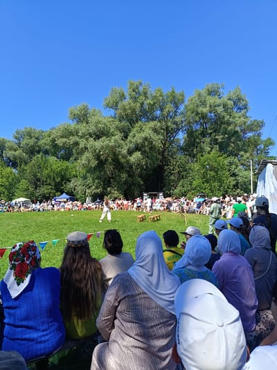 In the city of Tulbay, Russia, a daytime outdoor gathering takes place on a grassy field bordered by lush green trees. A crowd of people, seated on the ground or on benches, watches a performance. In the foreground, several individuals are seen from behind, some wearing headscarves and hats. In the mid-ground, a performer in white attire stands on the grass, facing a seated audience. Colorful triangular flags are strung across the field, adding a festive atmosphere to the scene. The sky above is a clear, vibrant blue.
