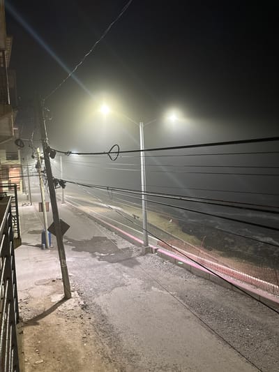 The image depicts a foggy night scene in Tijuana, Mexico. A street runs through the center of the frame, bordered by a sidewalk and a construction area with orange netting. Overhead, a complex network of power lines crisscrosses the sky. Streetlights illuminate the foggy environment, creating a hazy glow. A tall, leaning utility pole with numerous wires attached stands prominently on the left side of the street. The ground is a mix of asphalt and gravel, with some puddles on the sidewalk. The overall atmosphere is dark and somewhat desolate.