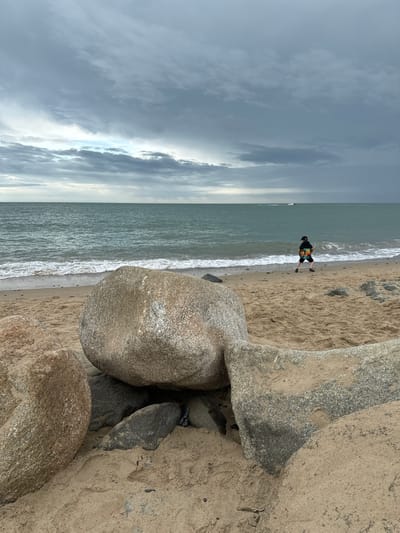 The image depicts a beach scene at Courtown, Ireland, under a heavily clouded sky. In the foreground, large grey and tan boulders are scattered on the sandy shore. The sea is a deep turquoise, with moderate waves breaking on the beach. A person, viewed from behind, wearing a dark cap, a colorful jacket with a tie-dye pattern, and dark shorts, stands on the wet sand near the water's edge, holding a bright orange object. In the distance, a small boat or jet ski is visible on the water. The overall atmosphere is one of a cool, overcast day at the seaside.