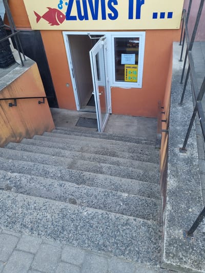 The image shows the entrance to a store called "Žuvis ir..." in Kuršėnai, Lithuania. The store's name is displayed in blue letters on a yellow sign above the entrance. To the left of the name is a red graphic of a fish with bubbles. The entrance is a set of stairs leading down to a glass door that is partially open. A sign is visible in the window to the right of the door, indicating the store's opening hours. The building has an orange facade. The stairs are made of concrete with small embedded stones. There are metal railings on the right side of the stairs. The environment appears to be outdoors, possibly on a sidewalk or street.