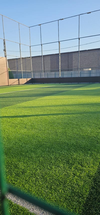 The image shows an outdoor artificial turf sports field under a clear blue sky. The field is green with a white line marking. The edges of the field are enclosed by a metal fence with netting, and there are floodlights attached to the top of the fence. The sun casts long shadows across the field, indicating it is either early morning or late afternoon. In the background, there is a brown wall and a dark fence with netting. There is no indication of the specific location within the city of Овощной, Russia. There are no people or actions depicted in the image.