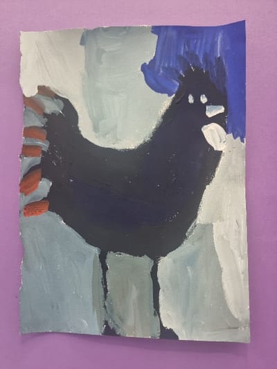 The image displays a vertically oriented, hand-painted artwork on paper, positioned against a uniform purple background. The artwork depicts a stylized dark blue bird, identifiable as a rooster or hen, facing right. Its body is dark blue, featuring an irregular crest of blue brushstrokes on its head, two small white circular eyes, and a prominent white beak or wattle. The bird's tail, visible on the left, is represented by several horizontal reddish-brown strokes. The background of the painting is rendered in broad, vertical brushstrokes of light blue, grey, and dark blue hues. The edges of the paper are irregular, particularly along the top and left sides, suggesting they may be hand-torn. This scene is located in Jaunmārupe, Latvia.