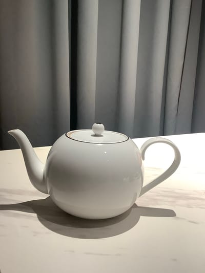 A white ceramic teapot with a curved handle and a spout is placed on a white surface. The teapot has a lid with a small knob and a thin dark line around the rim. The surface appears to be marble or a similar material with subtle veining. Behind the teapot, grey curtains are visible, creating a backdrop with vertical folds. The lighting creates shadows from the teapot on the surface. The scene is indoors, likely in a domestic or commercial setting, and there is no indication of the city of Nikul'skoye, Russia.