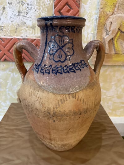 An aged, terracotta-colored vase with two handles and a dark blue rim is displayed on a light brown, textured surface. The vase features a lighter band around its neck, decorated with blue floral motifs and an inscription. The lower portion of the vase has incised linear patterns. In the background, there is a textured wall with faded artwork and decorative elements. The lighting suggests an indoor setting. The image is a close-up shot focused on the vase, with the surrounding environment serving as a backdrop. The specific location in the city of Fort-Şevtşenko, Kazakhstan is not visually identifiable from the image itself.