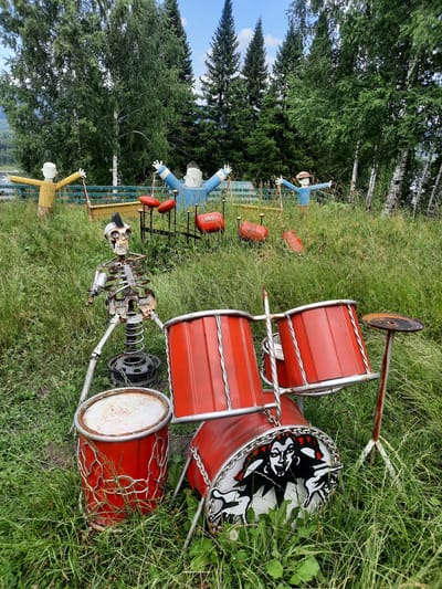 The image captures an outdoor scene featuring a unique assemblage of artistic installations in a grassy area surrounded by trees under a cloudy sky. In the foreground, a skeletal figure, constructed from scrap metal and springs, is positioned behind a red drum set. The drum set itself is made of red barrels with white rims and a decorative image of a jester on the bass drum. To the right of the drum set, a single cymbal is mounted on a metal stand.
In the background, several human-like figures, seemingly made from wood or other found materials, are arranged across the landscape. One figure, clad in blue and with arms outstretched, stands near a collection of red drums. Another figure, dressed in yellow, stands to the left. Further back, more figures and scattered drum components can be observed, suggesting a playful, perhaps musical, theme for these outdoor sculptures. The environment is characterized by tall, lush green grass and a backdrop of dense green trees, including birch trees with their distinctive white bark, and darker coniferous trees. The overall impression is one of an art installation set in a natural, overgrown setting. The location is identified as Зеленогорский, Russia.