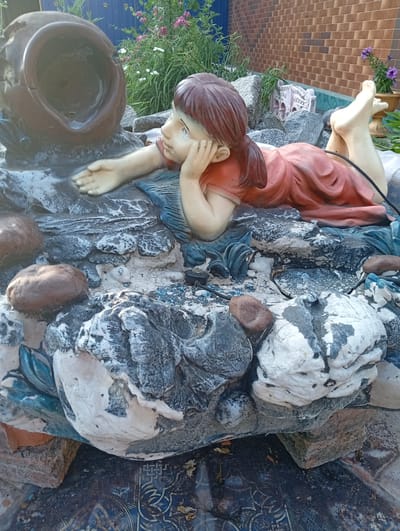 This media file features a garden ornament depicting a young girl in a red dress, lying on her side with her head propped on her hand. She appears to be looking towards a large, dark brown, pot-like object. The ornament is placed within a rock garden setting, with various stones and some foliage around it. In the background, a brick wall and a blue fence are visible, along with some flowering plants. The scene appears to be outdoors, likely during the daytime, given the natural lighting. The overall impression is one of a whimsical or decorative garden feature. There is no visible text in the image.
