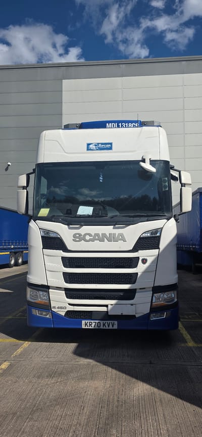 A white Scania R 450 truck with a blue lower front bumper and a license plate reading "KR70 KVV" is parked on a concrete surface marked with yellow lines. The truck has a "Borcan Transport Ltd" logo on the cab. In the background, a grey and white industrial building is visible under a bright blue sky with white clouds. To the left of the truck, a blue trailer is partially visible. The scene appears to be an outdoor area, likely a loading bay or parking lot associated with the industrial building. The truck is the primary subject and is facing the camera. There are no people visible in the image.