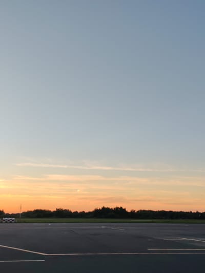 The image captures an outdoor scene at dusk, likely from the vicinity of Khrabrovo, Russia. A wide, dark asphalt surface, possibly a runway or taxiway, dominates the foreground. White lines mark the surface, suggesting a designated area for aircraft or vehicles. In the middle ground, a line of dense green trees forms a natural border against the horizon. Beyond the trees, the sky transitions from a pale, clear blue at the top to hues of orange and soft yellow near the horizon, indicating the presence of a sunset. A faint, circular shape, possibly the moon or a distant light, is visible in the upper portion of the orange sky. To the left, a red and white checkered marker or structure is partially visible, possibly a navigation aid or ground marker. The overall impression is one of an open, transitional space during the evening. There are no people or animals visible in the image.