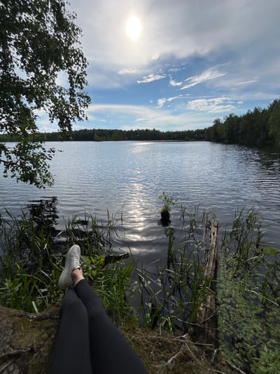A person's legs, clad in black leggings and white sneakers, are prominently featured in the foreground, resting on a moss-covered surface next to the dark water of a lake. Reeds and other aquatic plants grow along the water's edge, with a weathered wooden log partially submerged. The lake extends into the background, reflecting the bright sun and the sky, which is a mix of blue and white clouds. A line of dense green trees borders the far side of the lake, suggesting a natural, possibly rural, setting. The scene captures a moment of relaxation by the water under a bright, sunny sky. The specific location of Нагорный, Russia, is not discernible from the visual information present in the image.
