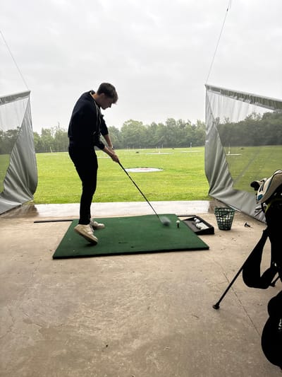 The image shows a young man in a black hoodie and black pants, swinging a golf club at a golf ball on a driving range at the Leyland Golf Centre in South Ribble, UK. He is standing on a green artificial turf mat. There are several golf balls scattered around the mat, including a white ball in flight, and a white golf tee is inserted into the turf. To the right of the man, there are several golf balls in a black plastic tray. A grey net enclosure covers the left side of the frame, and a grey net enclosure covers the right side of the frame. A golf bag is leaning against the right-side net enclosure. There is a grey bucket on the concrete ground behind the man, partially obscured by the net enclosure. The concrete ground is wet and appears to be rain-soaked. Trees and a grassy field are visible through the open areas of the net enclosures. The sky is cloudy and overcast. The image was likely taken on a rainy day.
