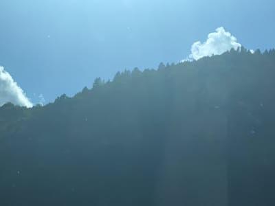 The image shows a hazy, sunlit sky with a large mountain slope covered in dark trees dominating the lower half. The sky is a bright blue with a few scattered clouds, one large, fluffy white cloud on the right side of the frame. Sunbeams are visible, cutting through the haze and illuminating the mountain's surface. The foreground is a dark, underexposed area, making it difficult to discern details. The scene is likely a natural landscape, possibly a view from a high vantage point looking down at a heavily forested mountain. There are no people or man-made structures visible in the image. The location is not explicitly indicated but the description of the landscape is consistent with the mountainous regions of Romania, including areas around Leordina.