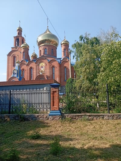 This is a medium-shot, eye-level outdoor photograph of a brick church with golden domes and blue accents, situated in Pokrov, Ukraine. The church is adorned with crosses, and one of its facades features a religious mosaic. The building is partially obscured by lush green trees and foliage. In the foreground, there is a black wrought-iron fence with a stone base. Beyond the fence, a patch of dry, yellowish grass with a few small green plants is visible. The sky is clear and blue, suggesting a sunny day. The time of day appears to be morning or early afternoon, indicated by the bright sunlight and distinct shadows. No people or specific activities are evident in the image. The architecture and the presence of the church strongly suggest a religious setting.